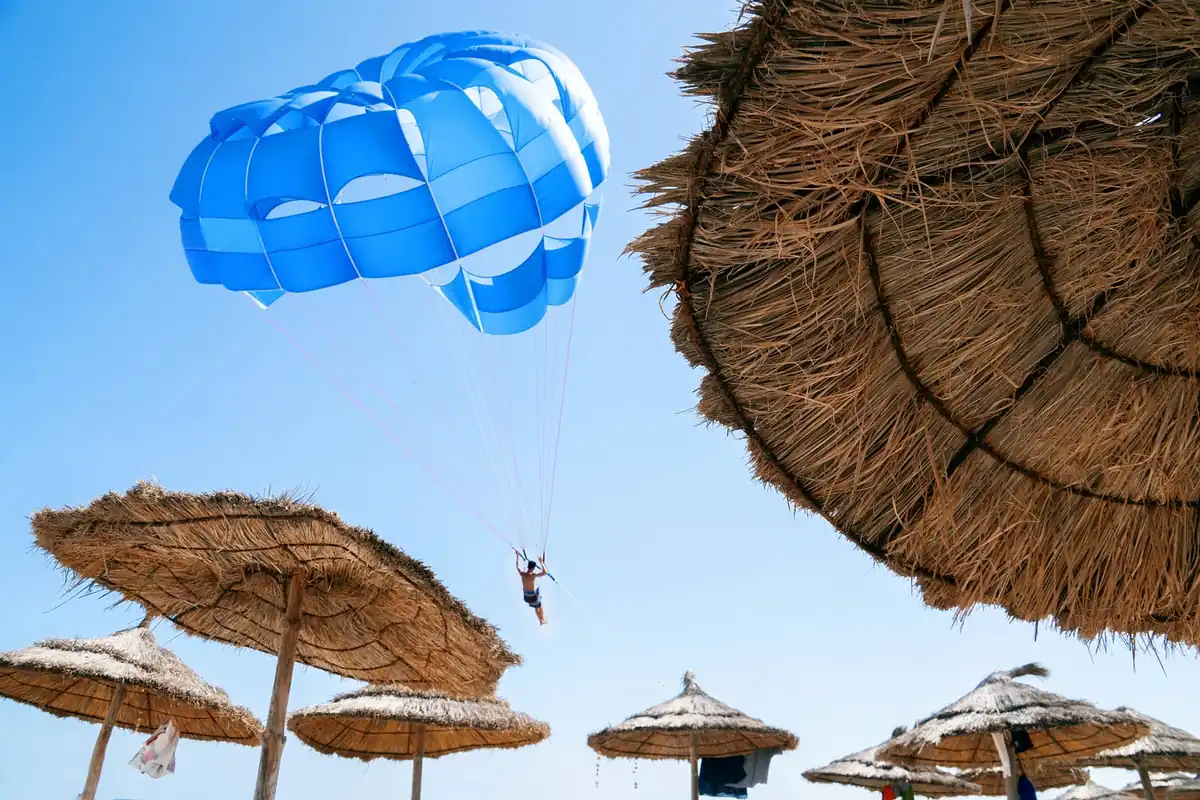 A blue parasail in the sky, seen between straw beach umbrellas