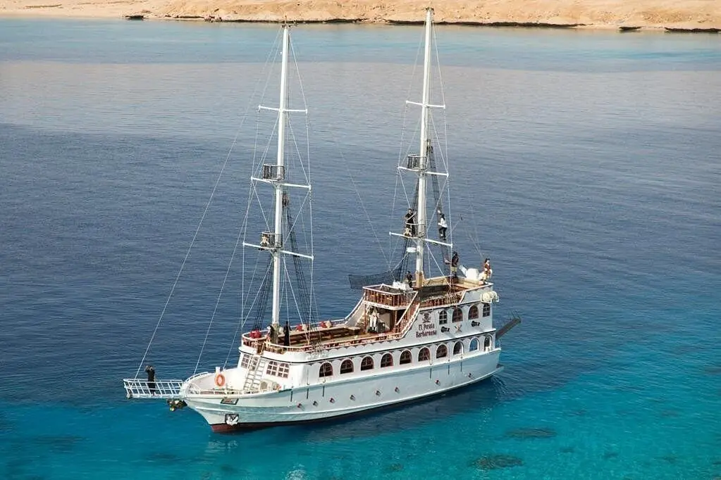 A two-masted boat at anchor in deep blue water, a sandy shore behind