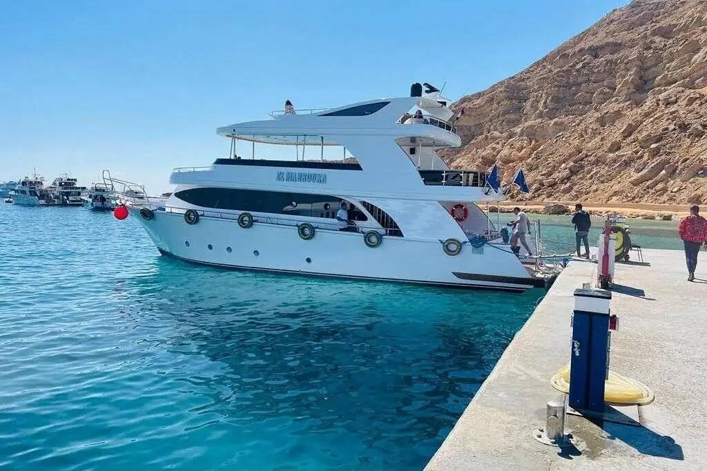 A white motor boat moored at a stone jetty, with a rocky mountain behind and more boats along the shore