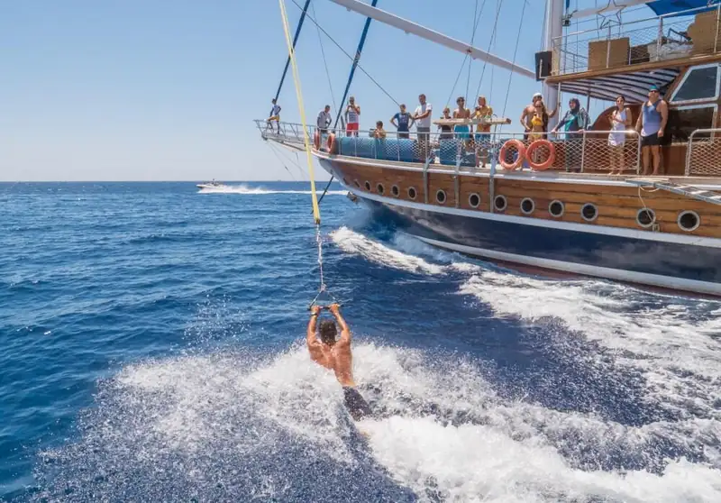 A guest swinging from a rope off the side of the wooden sailing yacht, skimming the wake while others watch from the deck