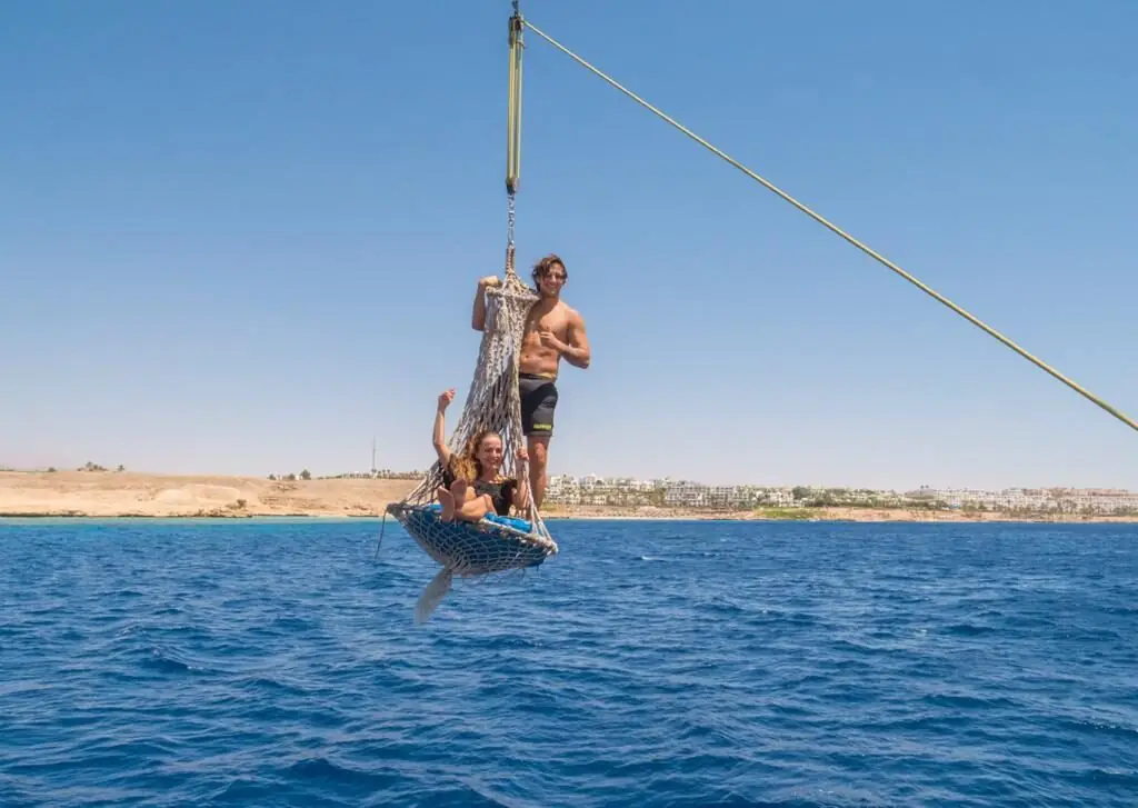 Two guests in a rope swing hanging from the yacht's rigging over the sea, the coast behind them