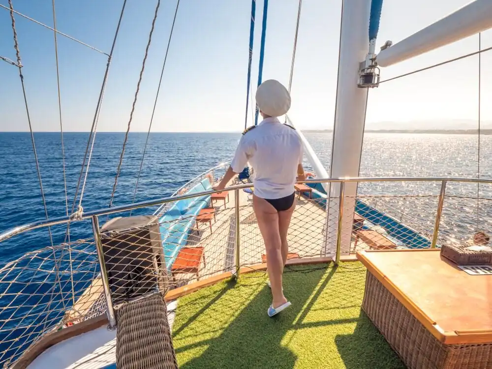 A guest in a white shirt and cap on the yacht's upper deck, looking out over the sea