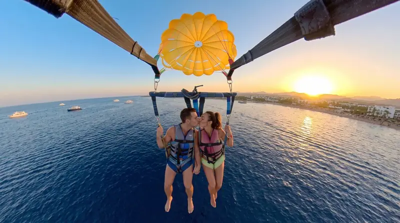 Two parasailers seen from above in their harness under a yellow canopy, the sun low over the bay