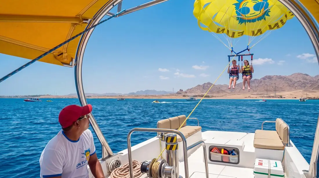 A boat driver in a red cap watching two parasailers behind the boat, the mountains on the horizon