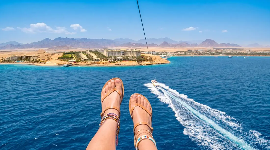 A parasailer's feet in sandals high above the blue bay, a speedboat's wake below and the coast behind