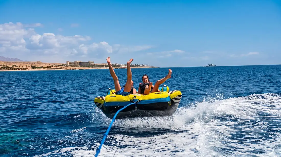 Two riders with their arms in the air on an inflatable tube, towed across the bay with the coast behind