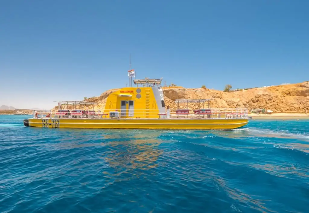 A yellow glass-bottom boat with an observation cabin on calm blue water, a sandy cliff behind